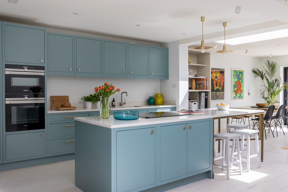 Transitional galley open plan kitchen in London with an undermount sink, flat-panel cabinets, blue cabinets, white splashback, black appliances, light hardwood floors, with island, beige floor and white benchtop.