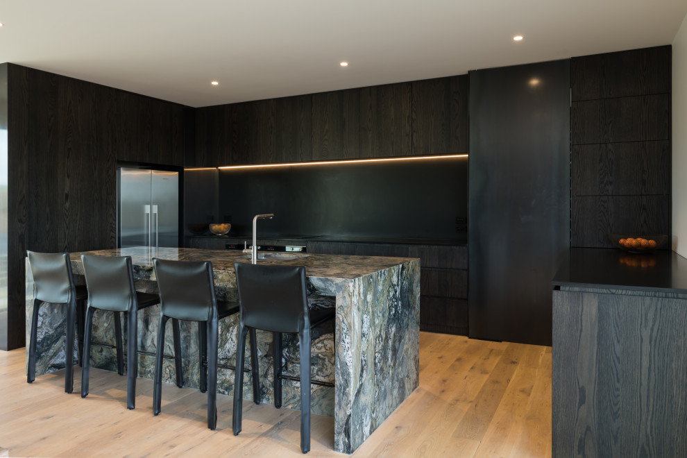 Mid-sized industrial u-shaped eat-in kitchen in Auckland with a single-bowl sink, flat-panel cabinets, dark wood cabinets, marble benchtops, black splashback, slate splashback, stainless steel appliances, light hardwood floors, with island, brown floor and black benchtop.