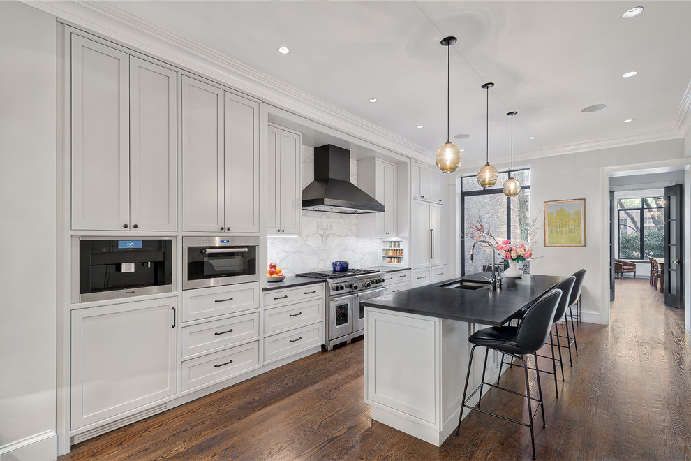 Inspiration for a mid-sized transitional galley open plan kitchen in New York with an undermount sink, white cabinets, soapstone benchtops, white splashback, marble splashback, medium hardwood floors, with island, brown floor and black benchtop.