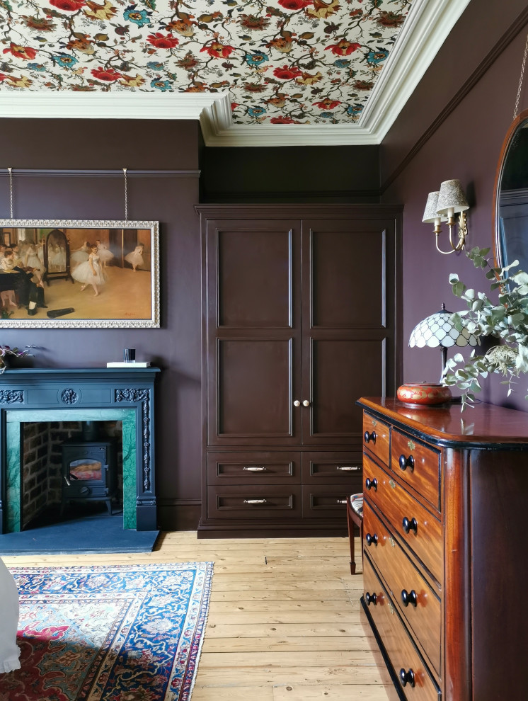 Modelo de dormitorio principal tradicional renovado grande con paredes púrpuras, suelo de madera en tonos medios, estufa de leña, marco de chimenea de piedra, suelo beige y papel pintado
