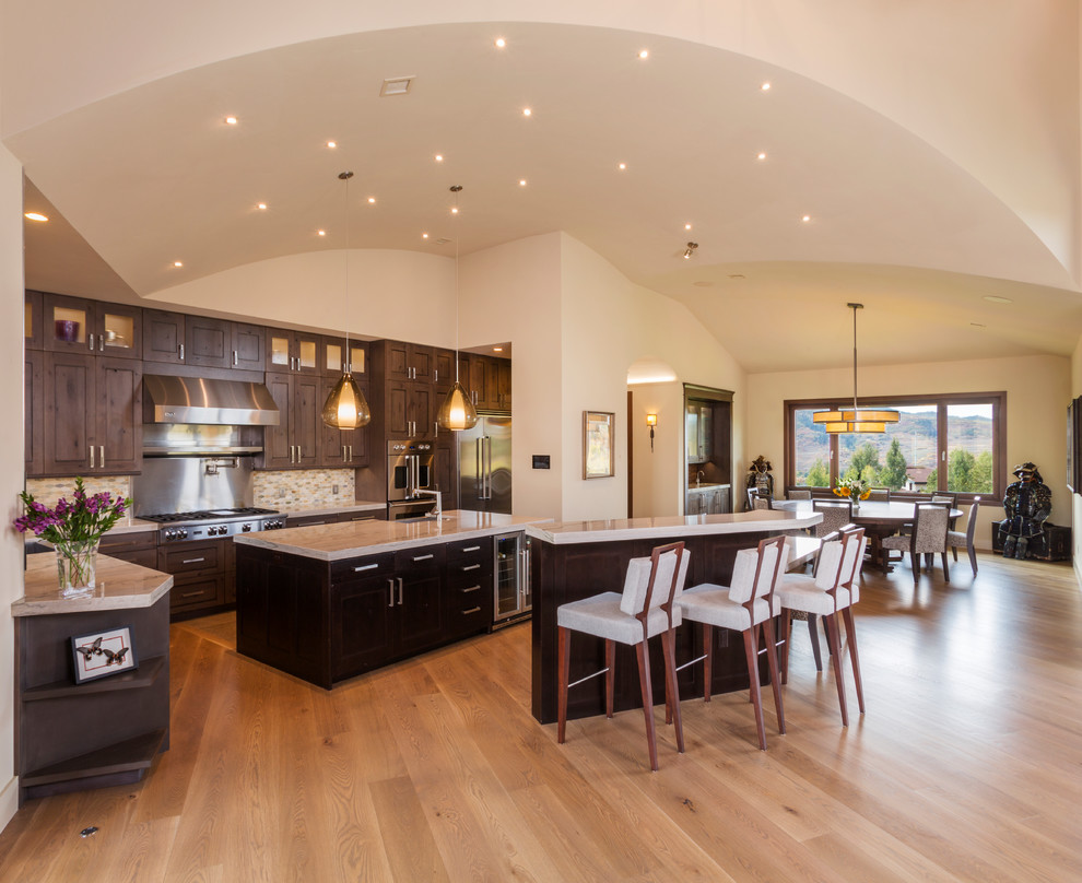 This is an example of a large contemporary u-shaped eat-in kitchen in Denver with a farmhouse sink, recessed-panel cabinets, dark wood cabinets, limestone benchtops, multi-coloured splashback, matchstick tile splashback, stainless steel appliances, light hardwood floors, multiple islands, beige floor and beige benchtop.
