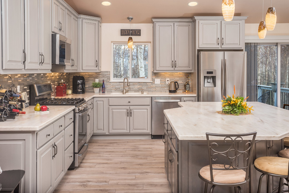 Mid-sized transitional l-shaped open concept kitchen photo with an undermount sink, raised-panel cabinets, gray cabinets, solid surface countertops, metallic backsplash, mosaic tile backsplash, stainless steel appliances and an island