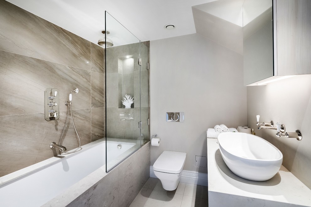 This is an example of a mid-sized contemporary kids bathroom in London with grey cabinets, an alcove tub, a shower/bathtub combo, a wall-mount toilet, gray tile, grey walls, a vessel sink, grey floor, a hinged shower door, white benchtops, a single vanity and a floating vanity.