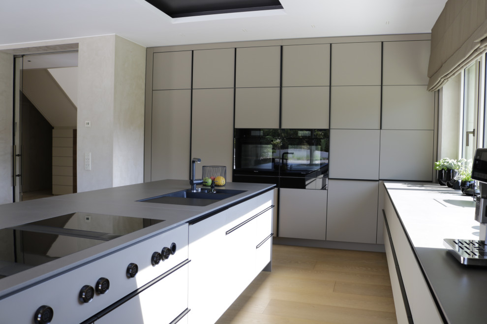 This is an example of an expansive modern grey and white galley open plan kitchen in Other with a built-in sink, flat-panel cabinets, grey cabinets, composite countertops, beige splashback, wood splashback, black appliances, medium hardwood flooring, an island, brown floors, grey worktops and a wallpapered ceiling.