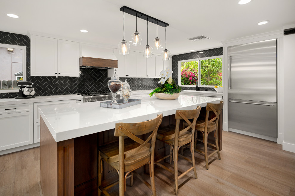 Inspiration for a mid-sized country l-shaped eat-in kitchen in Orange County with a farmhouse sink, shaker cabinets, white cabinets, quartz benchtops, blue splashback, ceramic splashback, stainless steel appliances, laminate floors and with island.