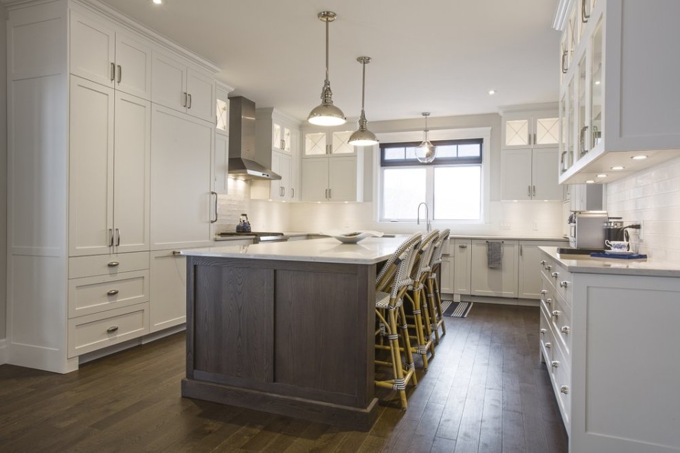 Large transitional u-shaped kitchen in New York with a farmhouse sink, shaker cabinets, white cabinets, solid surface benchtops, white splashback, subway tile splashback, panelled appliances, dark hardwood floors, with island and brown floor.