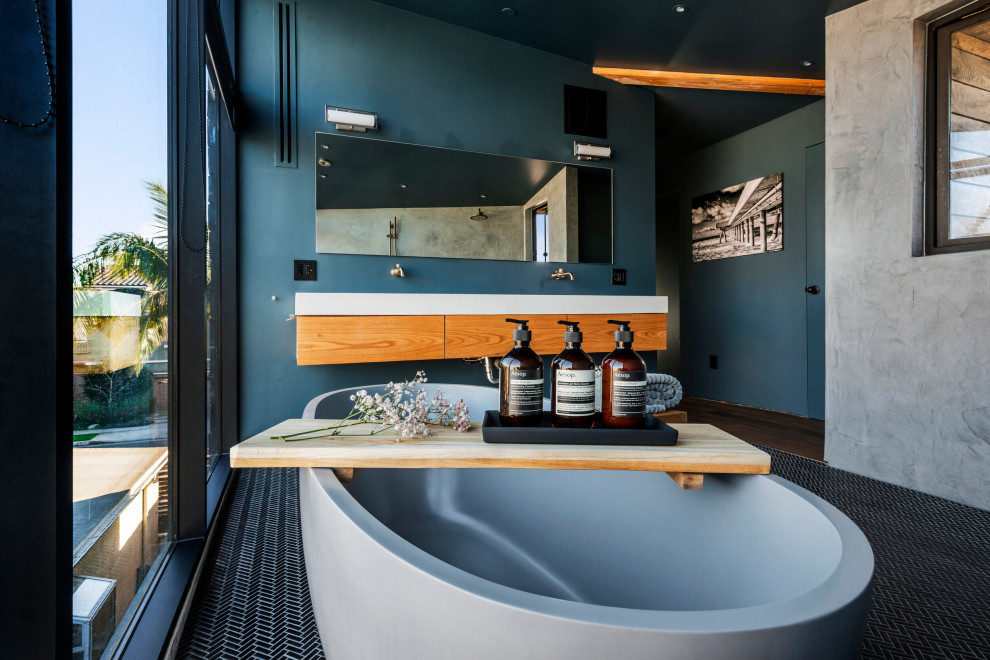 This is an example of a large industrial bathroom in Los Angeles with flat-panel cabinets, medium wood cabinets, a freestanding tub, blue walls, black floor, white benchtops, a single vanity, a floating vanity, porcelain floors and a niche.