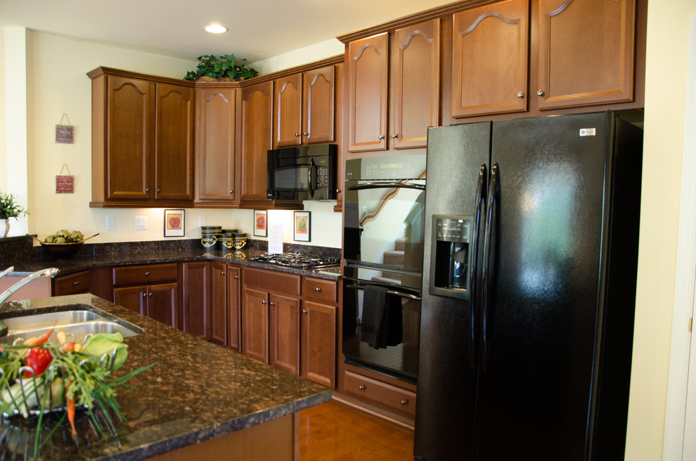 L-shaped medium tone wood floor eat-in kitchen photo with dark wood cabinets, granite countertops, beige backsplash, black appliances and an island
