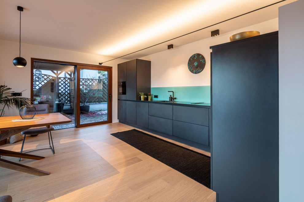 Photo of a large modern l-shaped eat-in kitchen in Other with a drop-in sink, flat-panel cabinets, grey cabinets, granite benchtops, green splashback, glass sheet splashback, black appliances, painted wood floors, no island, brown floor and black benchtop.