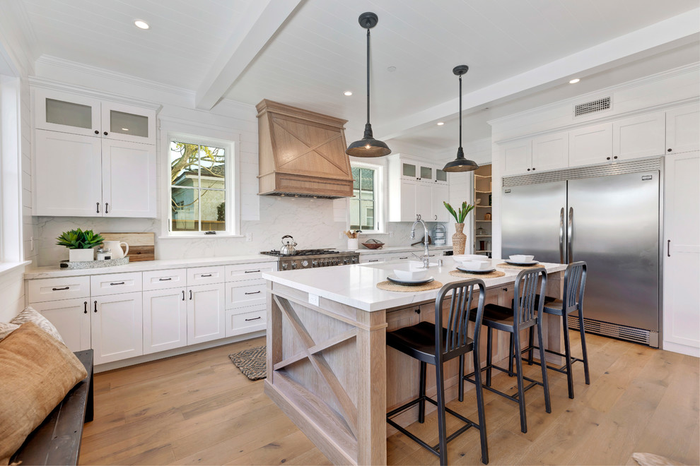 Design ideas for a country l-shaped kitchen in Los Angeles with an undermount sink, shaker cabinets, white cabinets, white splashback, stainless steel appliances, medium hardwood floors, with island, brown floor and white benchtop.