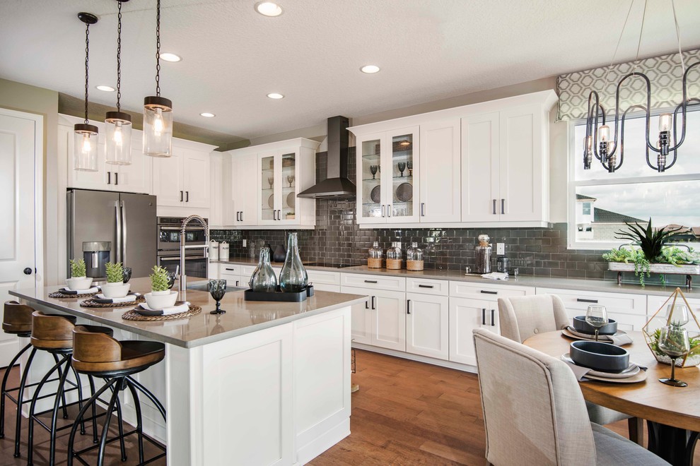 This is an example of a country l-shaped eat-in kitchen in Orlando with a drop-in sink, recessed-panel cabinets, white cabinets, quartzite benchtops, grey splashback, glass tile splashback, stainless steel appliances, medium hardwood floors, with island, brown floor and grey benchtop.