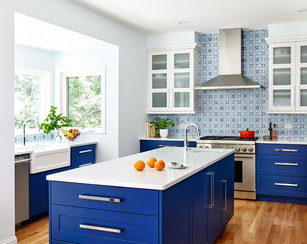 Photo of a transitional kitchen in DC Metro with a farmhouse sink, shaker cabinets, blue cabinets, multi-coloured splashback, stainless steel appliances, light hardwood floors, with island and white benchtop.