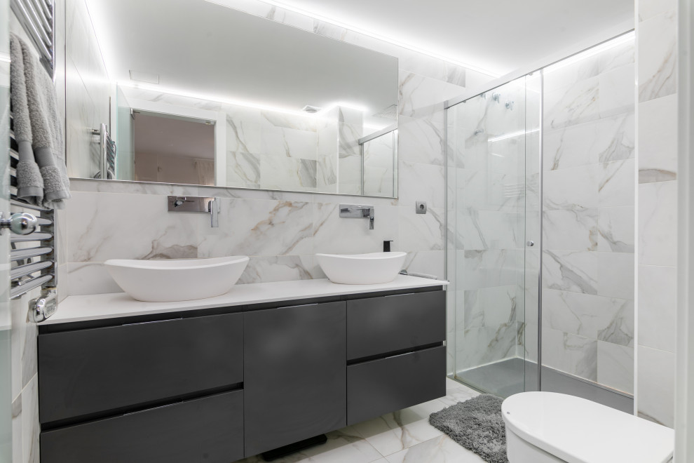 This is an example of a mid-sized contemporary master bathroom in Barcelona with flat-panel cabinets, grey cabinets, a curbless shower, a one-piece toilet, multi-coloured tile, ceramic tile, multi-coloured walls, ceramic floors, a drop-in sink, limestone benchtops, multi-coloured floor, a sliding shower screen, white benchtops, a double vanity and a floating vanity.