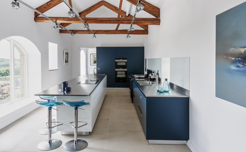 Inspiration for a contemporary l-shaped kitchen in London with an integrated sink, flat-panel cabinets, blue cabinets, stainless steel benchtops, white splashback, black appliances, with island, beige floor, grey benchtop, exposed beam and vaulted.