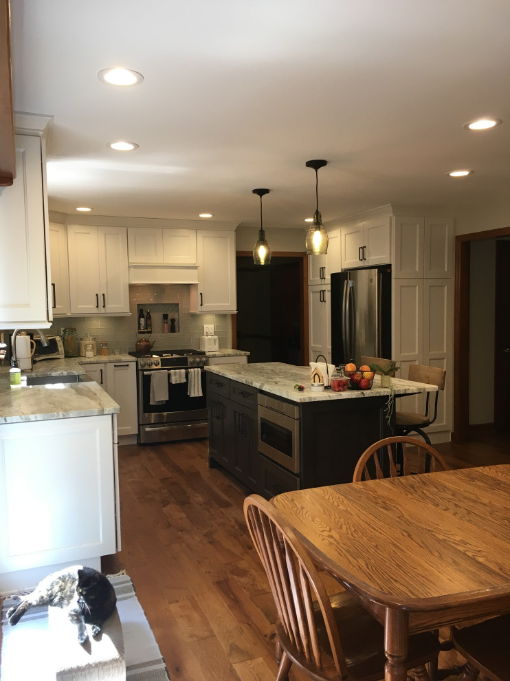 Inspiration for a mid-sized transitional u-shaped medium tone wood floor and brown floor eat-in kitchen remodel with a farmhouse sink, recessed-panel cabinets, white cabinets, granite countertops, gray backsplash, glass tile backsplash, stainless steel appliances, an island and gray countertops