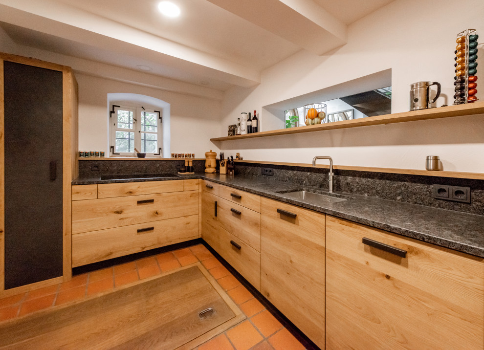 This is an example of a mid-sized country l-shaped separate kitchen in Other with a single-bowl sink, flat-panel cabinets, medium wood cabinets, granite benchtops, grey splashback, granite splashback, black appliances, terra-cotta floors, no island, red floor and grey benchtop.