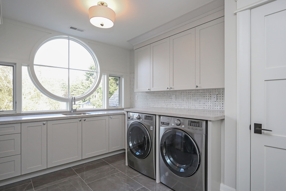 This is an example of a large transitional l-shaped dedicated laundry room in Toronto with an undermount sink, shaker cabinets, grey cabinets, quartz benchtops, grey walls, travertine floors, a side-by-side washer and dryer and grey floor.