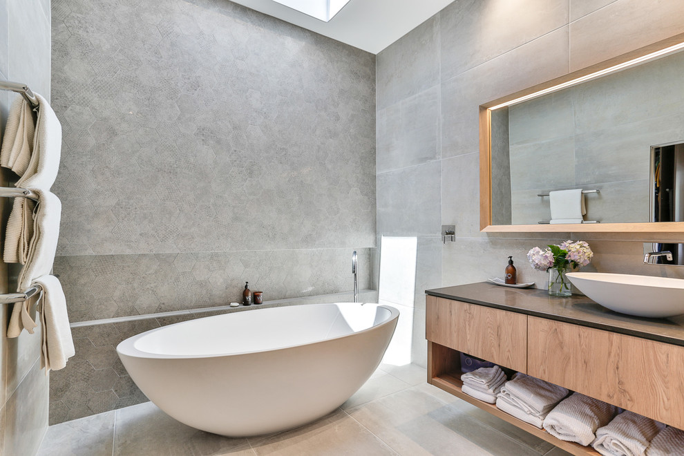 Mid-sized contemporary 3/4 bathroom in Auckland with light wood cabinets, a freestanding tub, gray tile, ceramic tile, grey walls, engineered quartz benchtops, black benchtops, flat-panel cabinets, a vessel sink and grey floor.