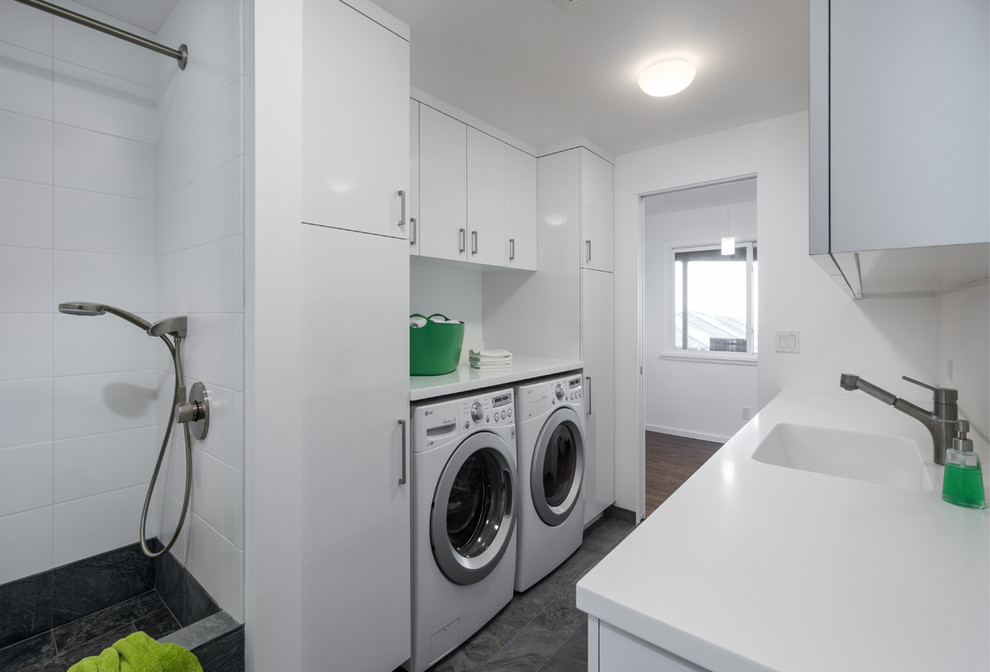 Design ideas for a mid-sized contemporary galley utility room in Portland with flat-panel cabinets, white cabinets, white walls, a side-by-side washer and dryer, solid surface benchtops, an integrated sink, white splashback, slate floors, grey floor and white benchtop.