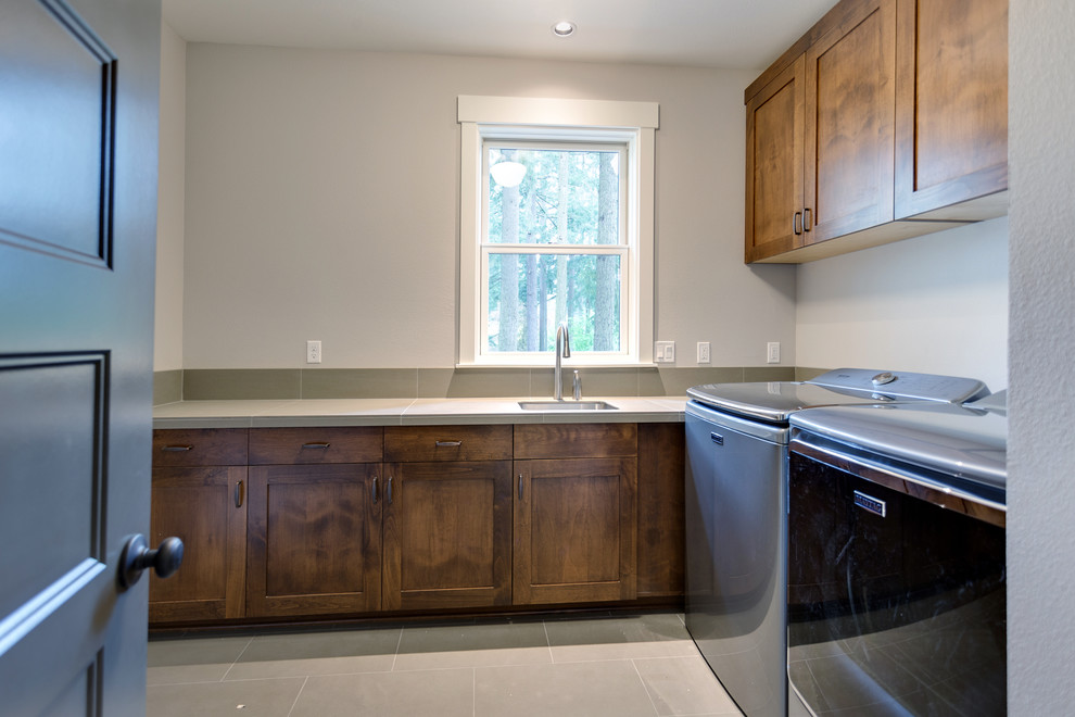 This is an example of a country l-shaped dedicated laundry room in Salt Lake City with an undermount sink, recessed-panel cabinets, dark wood cabinets, tile benchtops, grey walls, porcelain floors, a side-by-side washer and dryer, grey floor and grey benchtop.