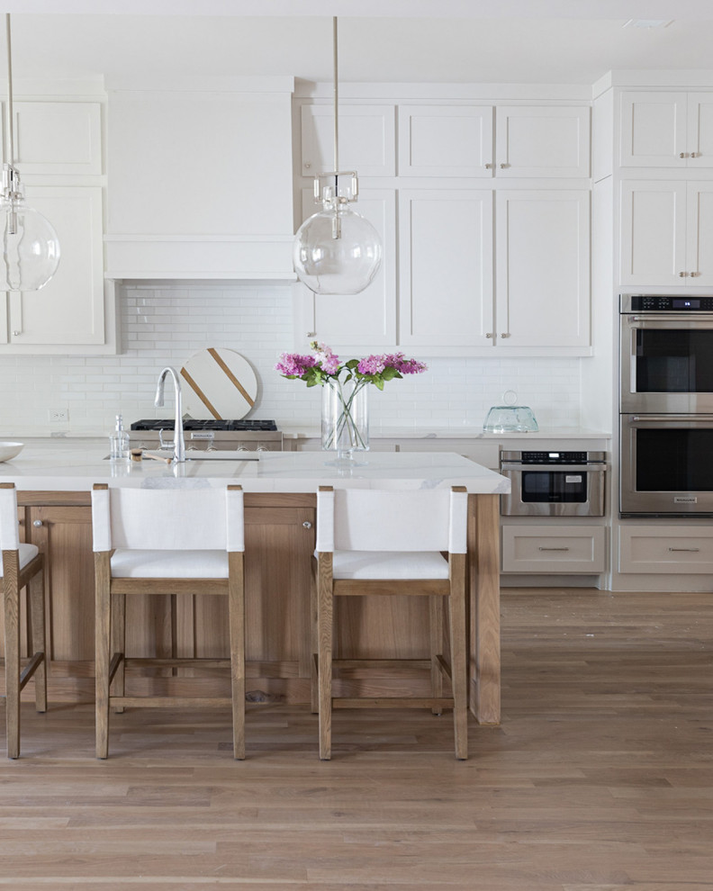 Photo of an expansive coastal single-wall kitchen/diner in Dallas with a submerged sink, shaker cabinets, white cabinets, engineered stone countertops, white splashback, porcelain splashback, stainless steel appliances, light hardwood flooring, an island, brown floors and white worktops.