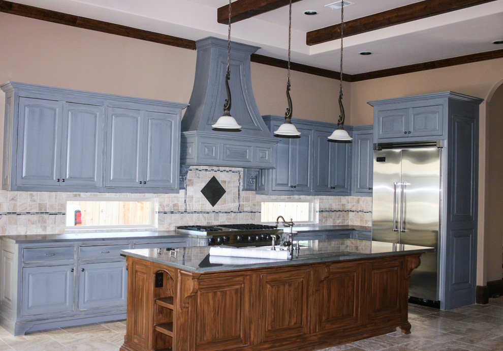 Photo of a large traditional l-shaped eat-in kitchen in Houston with an undermount sink, raised-panel cabinets, distressed cabinets, soapstone benchtops, beige splashback, stone tile splashback, stainless steel appliances, limestone floors and with island.