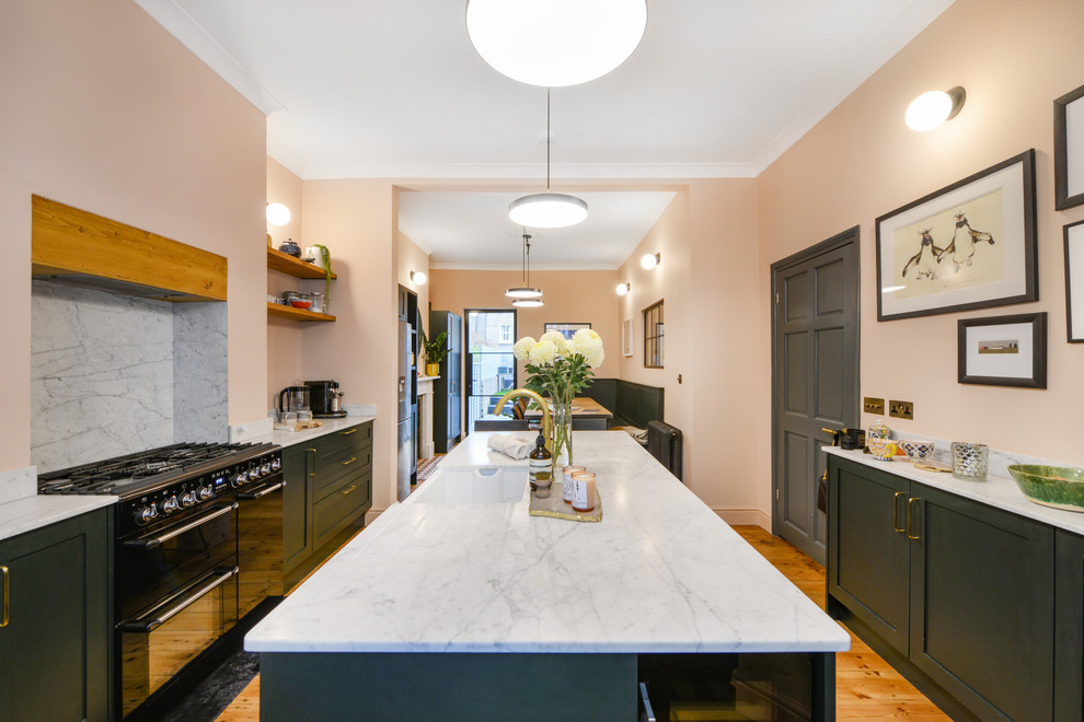 This is an example of a scandinavian galley eat-in kitchen in London with a farmhouse sink, shaker cabinets, green cabinets, grey splashback, black appliances, medium hardwood floors, with island, orange floor and white benchtop.