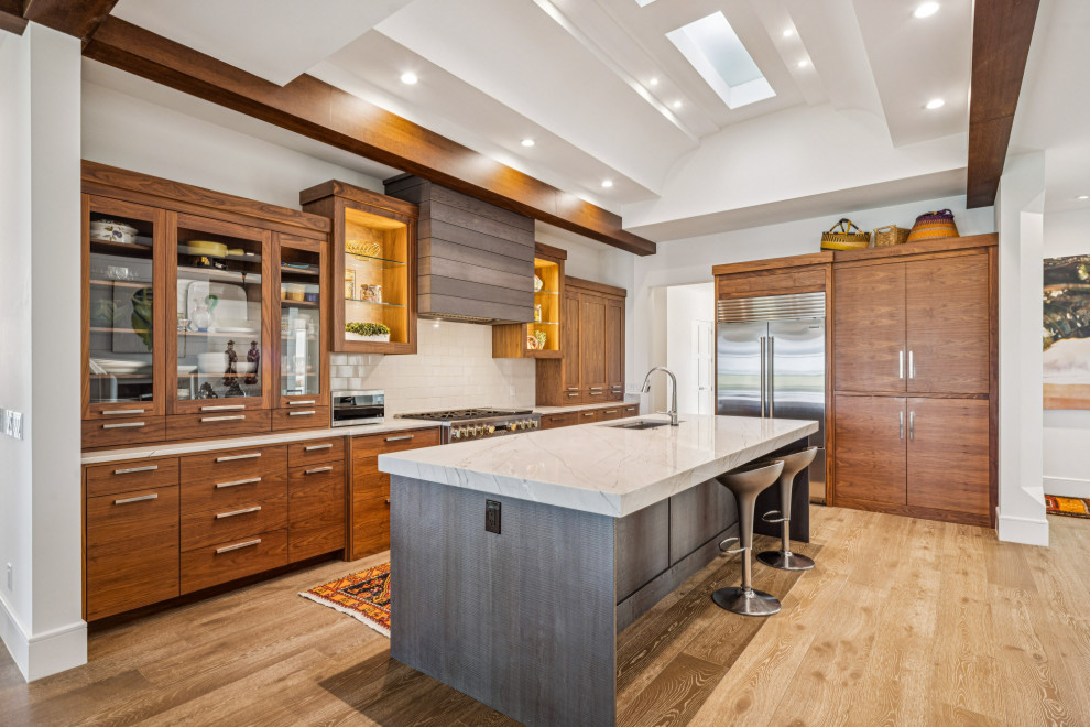 Inspiration for a transitional l-shaped light wood floor, beige floor, exposed beam and vaulted ceiling kitchen remodel with an undermount sink, shaker cabinets, medium tone wood cabinets, white backsplash, stone slab backsplash, stainless steel appliances, an island and white countertops
