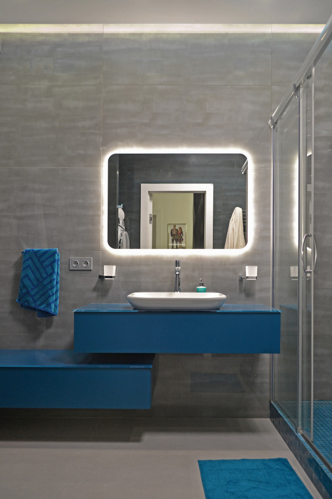 На фото: ванная комната среднего размера в современном стиле с плоскими фасадами, синими фасадами, угловым душем, инсталляцией, серой плиткой, керамогранитной плиткой, синими стенами, полом из керамогранита, душевой кабиной, настольной раковиной, стеклянной столешницей, серым полом, душем с раздвижными дверями, синей столешницей, тумбой под одну раковину, подвесной тумбой и многоуровневым потолком