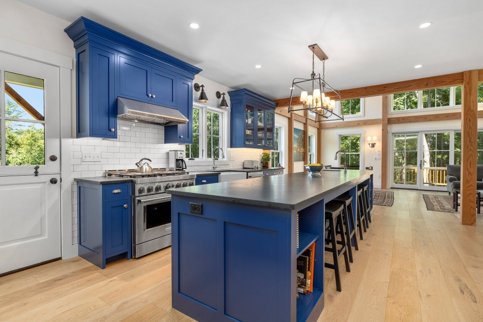 Mid-sized country single-wall eat-in kitchen in Boston with a farmhouse sink, shaker cabinets, blue cabinets, soapstone benchtops, white splashback, subway tile splashback, stainless steel appliances, light hardwood floors, with island and black benchtop.