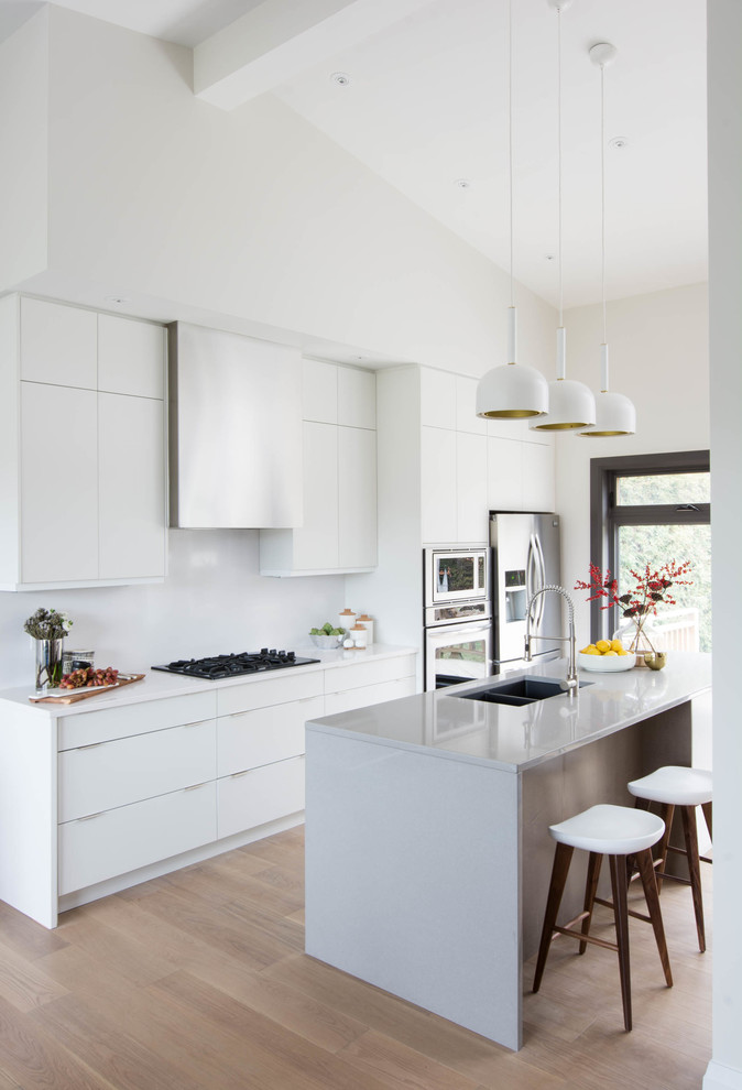 Photo of a mid-sized scandinavian single-wall open plan kitchen in Toronto with an undermount sink, flat-panel cabinets, white cabinets, quartzite benchtops, white splashback, stone slab splashback, stainless steel appliances, light hardwood floors and with island.