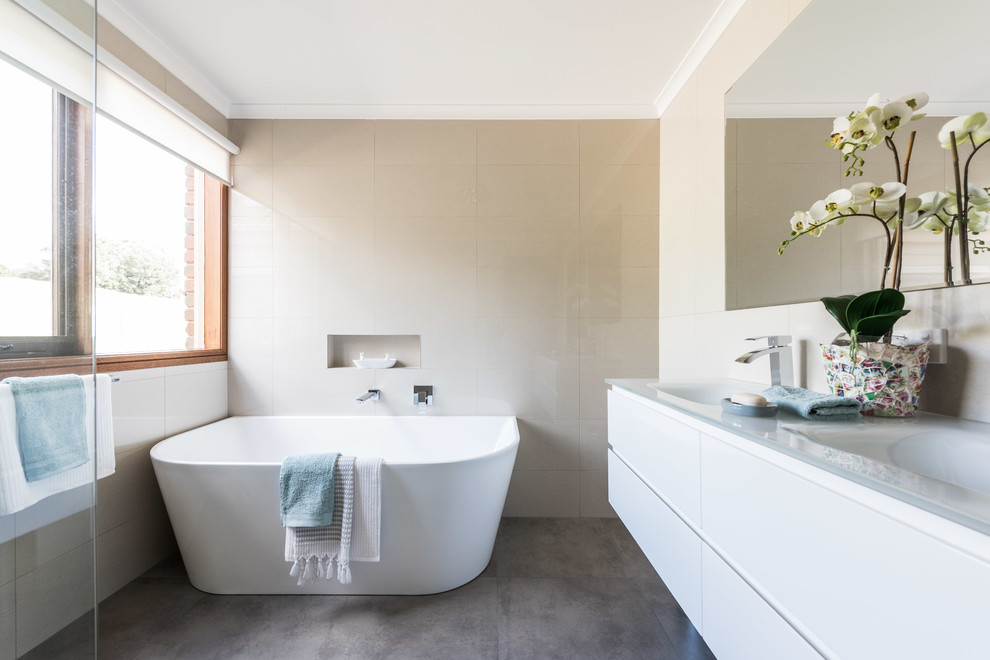 Small contemporary bathroom in Melbourne with white cabinets, a freestanding tub, beige tile, porcelain tile, porcelain floors, glass benchtops, grey floor, white benchtops, flat-panel cabinets and an integrated sink.