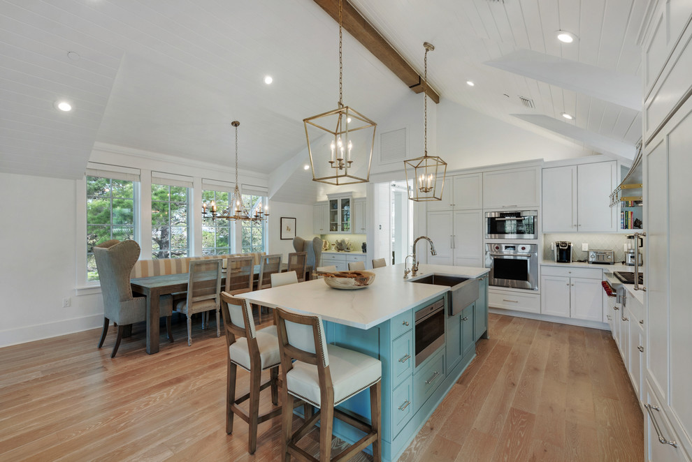 Photo of a beach style l-shaped kitchen in Miami with a farmhouse sink, shaker cabinets, white cabinets, panelled appliances, medium hardwood floors, with island, brown floor and white benchtop.