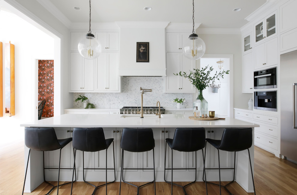 This is an example of a transitional l-shaped kitchen in Nashville with an undermount sink, shaker cabinets, white cabinets, white splashback, marble splashback, stainless steel appliances, medium hardwood floors, with island and brown floor.