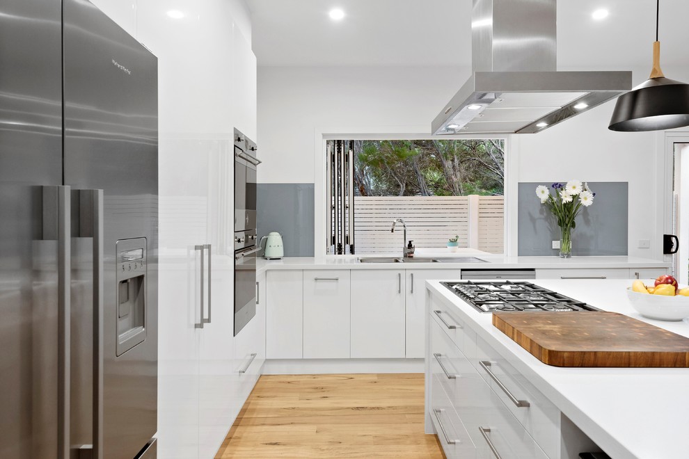 Design ideas for a beach style l-shaped kitchen in Melbourne with a drop-in sink, flat-panel cabinets, white cabinets, grey splashback, glass sheet splashback, stainless steel appliances, light hardwood floors, with island, beige floor and white benchtop.