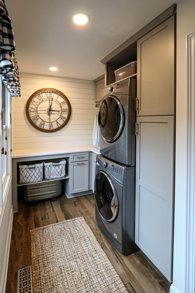Photo of a small transitional u-shaped dedicated laundry room in Cleveland with recessed-panel cabinets, grey cabinets, quartz benchtops, white splashback, shiplap splashback, grey walls, a stacked washer and dryer and white benchtop.