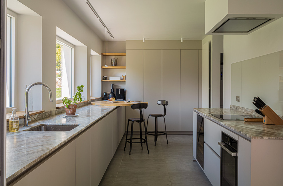 Photo of a large beach style u-shaped open plan kitchen in Nice with an undermount sink, flat-panel cabinets, beige cabinets, grey splashback, black appliances, ceramic floors, no island, beige floor and grey benchtop.