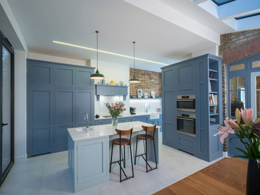 This is an example of a medium sized classic galley kitchen/diner in London with a belfast sink, shaker cabinets, blue cabinets, quartz worktops, white splashback, engineered quartz splashback, stainless steel appliances, porcelain flooring, an island, grey floors, white worktops and feature lighting.