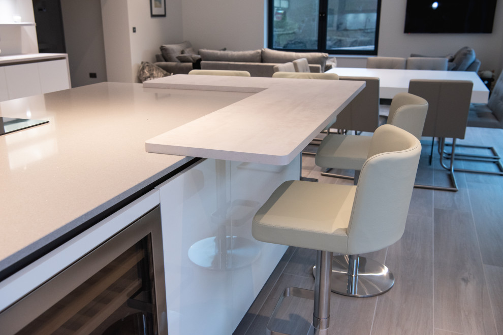 This is an example of a large modern l-shaped open plan kitchen in Kent with an undermount sink, flat-panel cabinets, white cabinets, quartzite benchtops, white splashback, glass sheet splashback, black appliances, light hardwood floors, with island and white benchtop.
