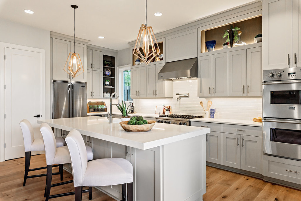 Design ideas for a beach style l-shaped kitchen in Portland with shaker cabinets, grey cabinets, white splashback, subway tile splashback, stainless steel appliances, light hardwood floors, with island and white benchtop.