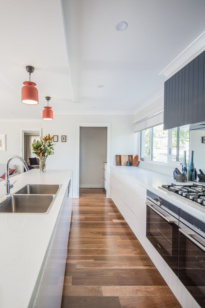 Photo of a large contemporary galley open plan kitchen in Brisbane with a double-bowl sink, white cabinets, quartz benchtops, mirror splashback, stainless steel appliances, medium hardwood floors, with island, brown floor and white benchtop.