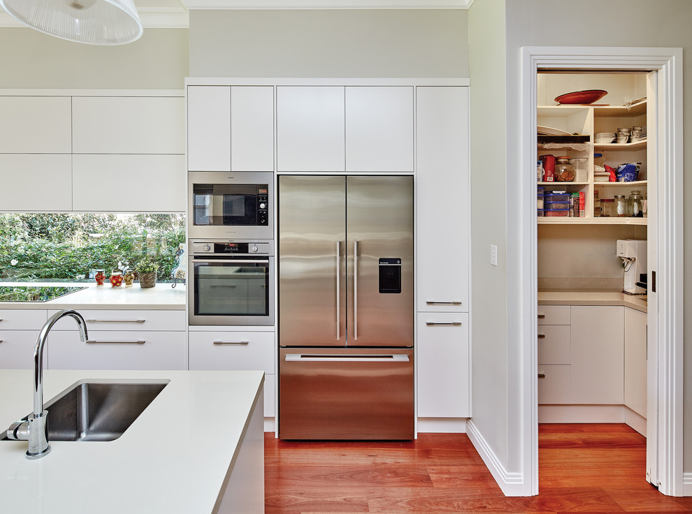 Photo of a large modern l-shaped kitchen pantry in Sydney with an integrated sink, flat-panel cabinets, white cabinets, quartz benchtops, window splashback, stainless steel appliances, medium hardwood floors, with island and brown floor.