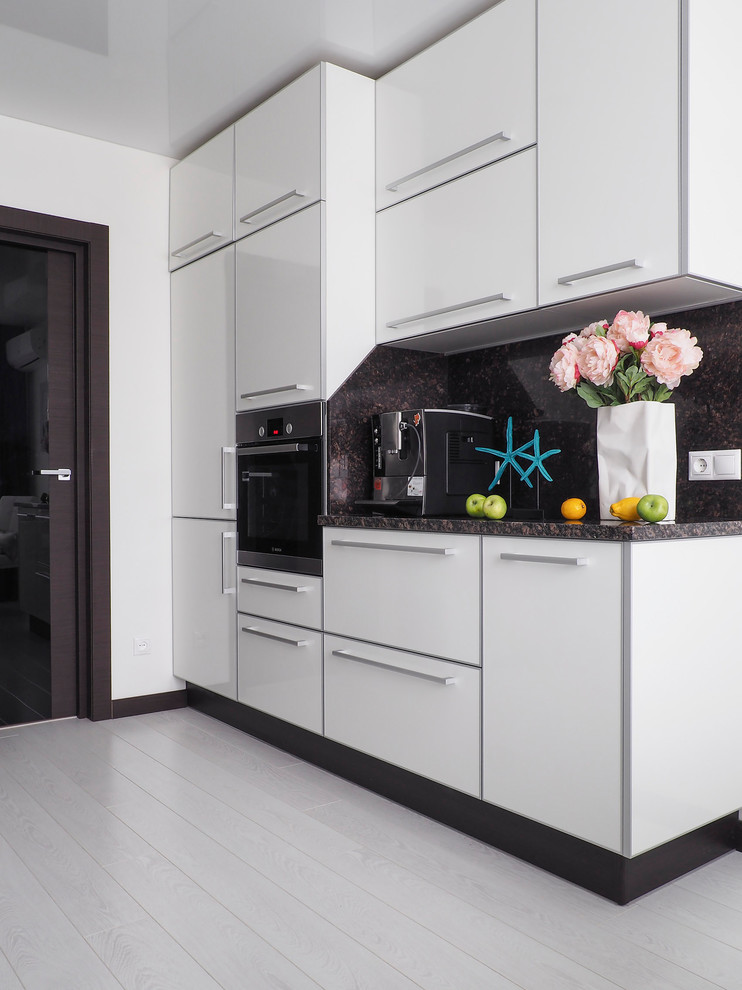 Contemporary single-wall eat-in kitchen in Other with flat-panel cabinets, white cabinets, black splashback, black appliances and white floor.