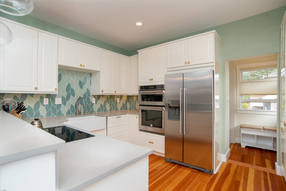 This is an example of a small beach style u-shaped eat-in kitchen in Seattle with a farmhouse sink, raised-panel cabinets, white cabinets, green splashback, ceramic splashback, stainless steel appliances, medium hardwood floors, no island and beige benchtop.