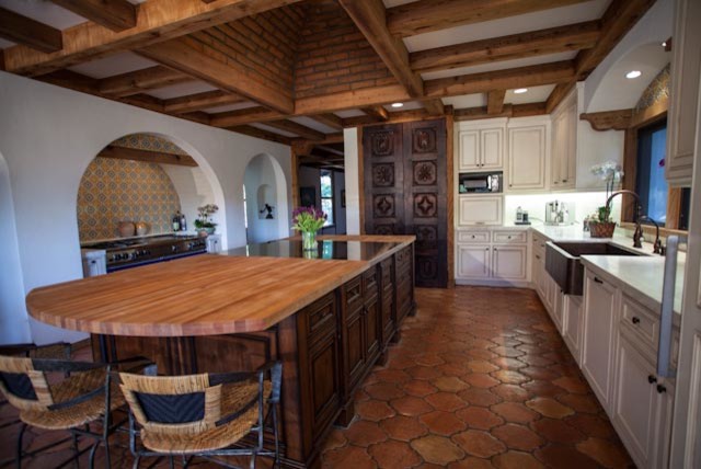 Photo of a mediterranean u-shaped eat-in kitchen in San Diego with an undermount sink, beaded inset cabinets, medium wood cabinets, wood benchtops, white splashback, stone tile splashback, black appliances and ceramic floors.