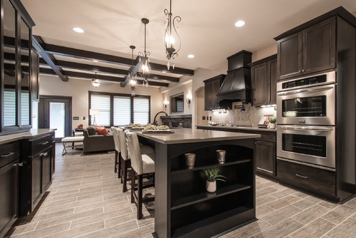 Warm transitional kitchen featuring dark wood kitchen cabinets, black range hood, stainless double ovens, a quartz-topped island with open shelves, and a beamed ceiling with pendant chandeliers.