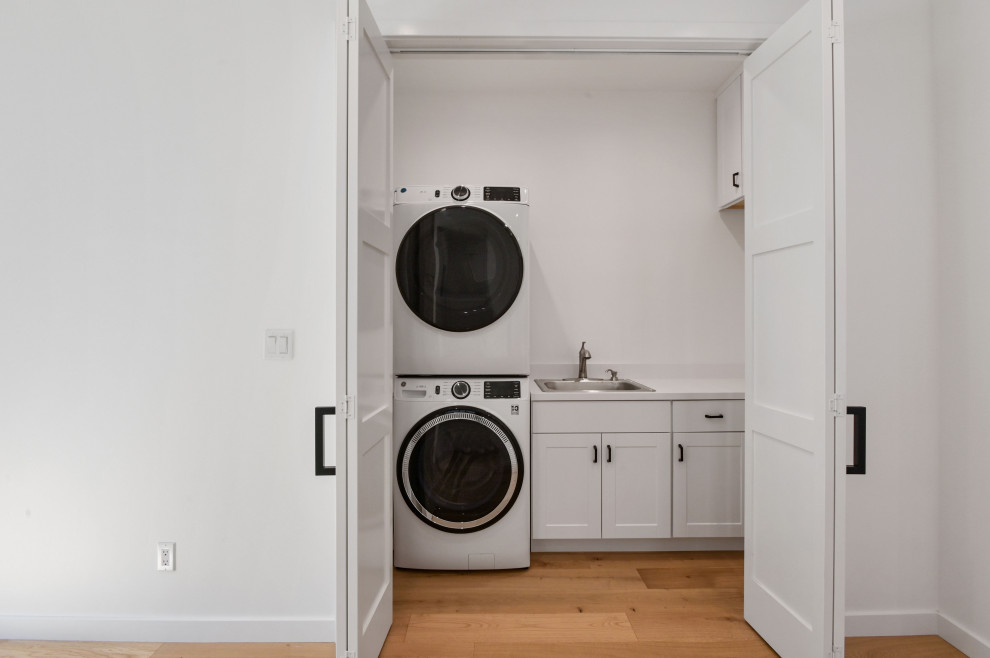 This is an example of a mid-sized contemporary single-wall laundry cupboard in San Francisco with a single-bowl sink, shaker cabinets, white cabinets, quartz benchtops, white splashback, engineered quartz splashback, white walls, light hardwood floors, a stacked washer and dryer and white benchtop.