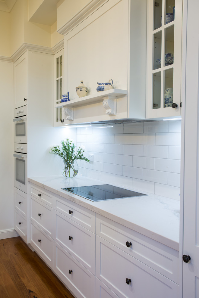 Large traditional galley kitchen pantry in Brisbane with a farmhouse sink, shaker cabinets, white cabinets, quartz benchtops, white splashback, subway tile splashback, white appliances, medium hardwood floors, with island, brown floor and white benchtop.