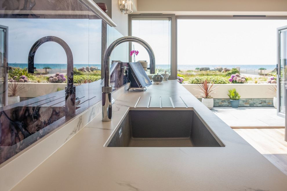 This is an example of a modern open plan kitchen in Sussex with a submerged sink, flat-panel cabinets, grey cabinets, glass sheet splashback, black appliances, an island and white worktops.