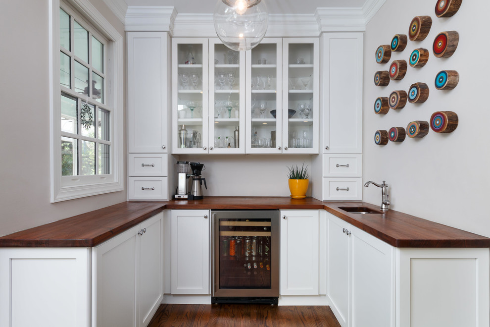 This is an example of a transitional u-shaped wet bar in Raleigh with an undermount sink, glass-front cabinets, white cabinets, wood benchtops, dark hardwood floors and brown floor.